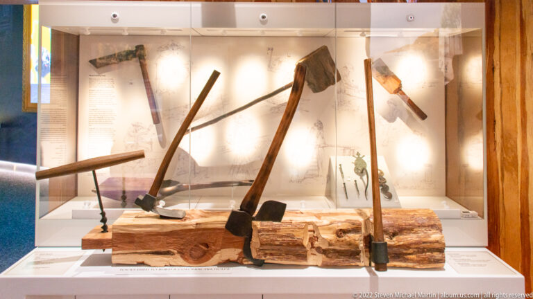 Long-handled tools and metal implements are displayed behind glass above rough timber sections.
