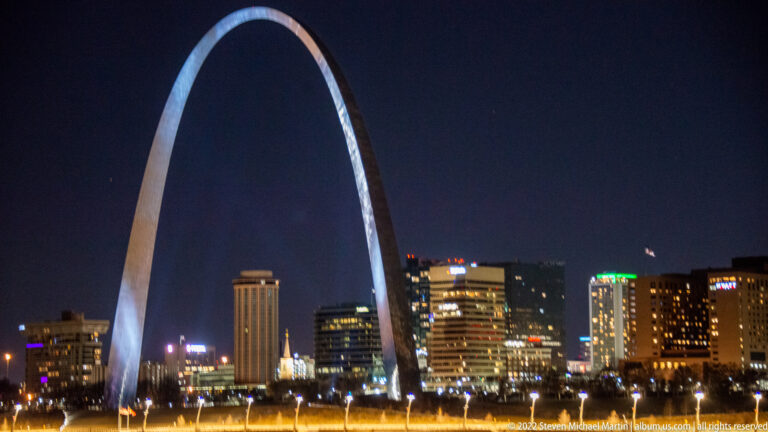 The full Gateway Arch rises over illuminated downtown buildings and park grounds at night.