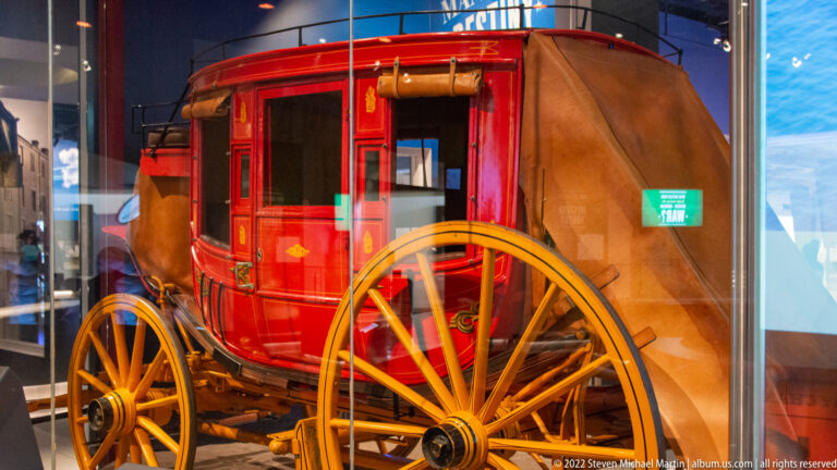 A red covered wagon with a canvas body and large yellow-spoked wheels stands behind glass.