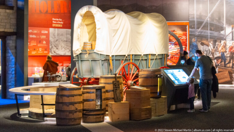 A canvas-covered wagon stands among barrels and crates as visitors view an interactive screen in the gallery.