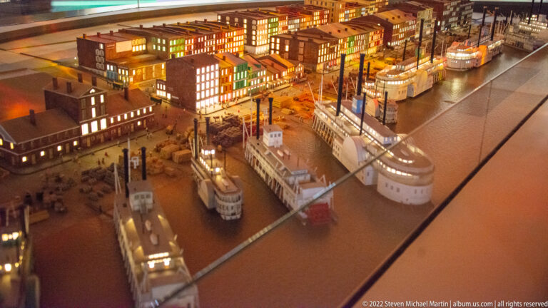 An illuminated scale model shows dense waterfront buildings, streets, and boats along a broad river channel.