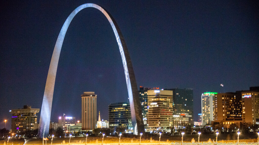 A broad night view shows the Gateway Arch above downtown towers and a brightly lit foreground.
