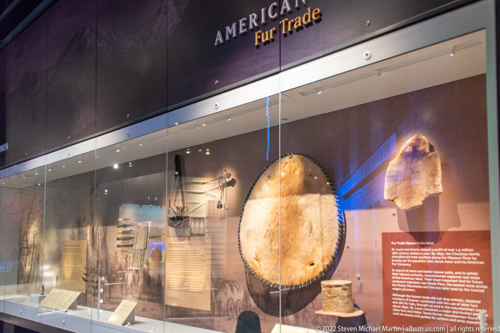 A large circular hide-like object and a smaller furred piece are displayed in a glass case beneath an “American Fur Trade” heading.