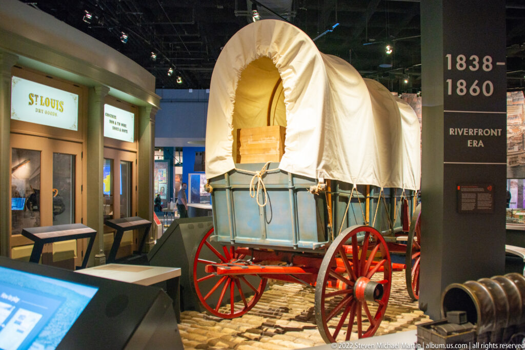 A canvas-covered wagon with red wheels stands among barrels and crates beside an interactive exhibit.