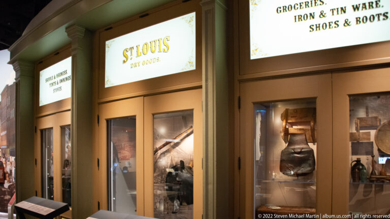 Storefronts labeled “St. Louis Dry Goods” and “Groceries, Iron & Tin Ware, Shoes & Boots” display goods behind glass.