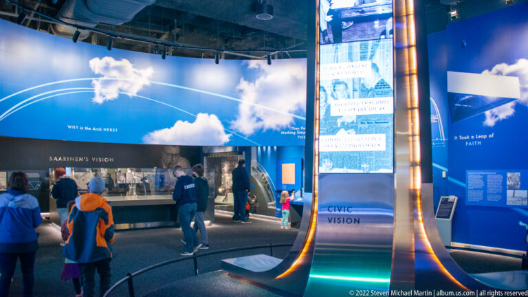 Visitors stand in a blue-lit gallery beside an arch-shaped structure and a panel labeled “Civic Vision.”