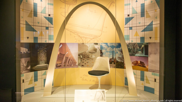 A white pedestal chair sits inside an arch-shaped frame before a wall of photographs and geometric panels.