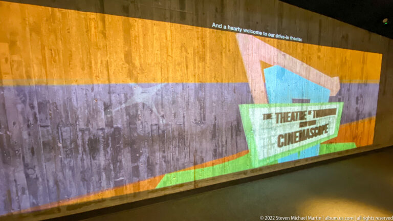 A colorful wall graphic with theater and cinema labels runs along a dim interior gallery.
