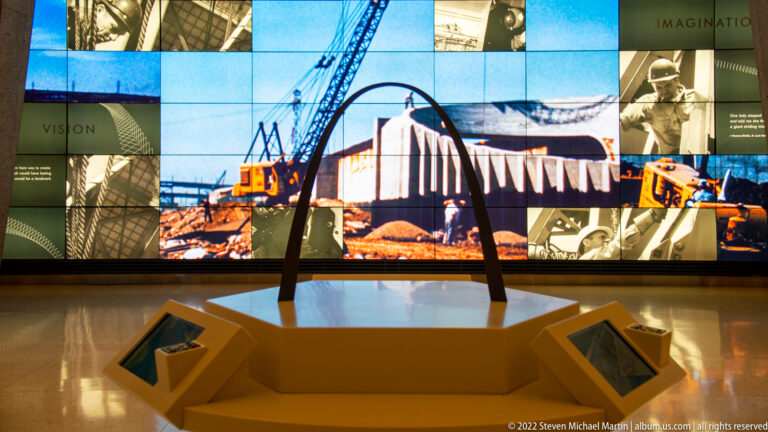 A small Gateway Arch model stands on a pedestal before a large screen of construction photographs and graphics.