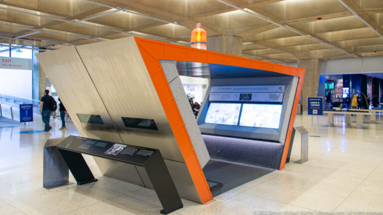 An orange-framed interactive exhibit with angled panels and screens stands in a spacious concrete museum hall.
