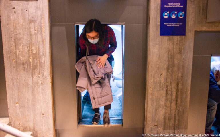 A visitor carrying a coat bends through a narrow restroom doorway framed by concrete partitions.