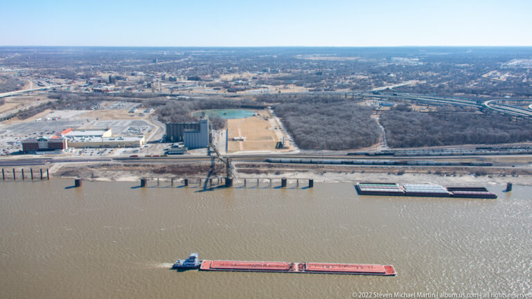 A red barge and smaller vessel move along the Mississippi River beside the St. Louis riverfront.