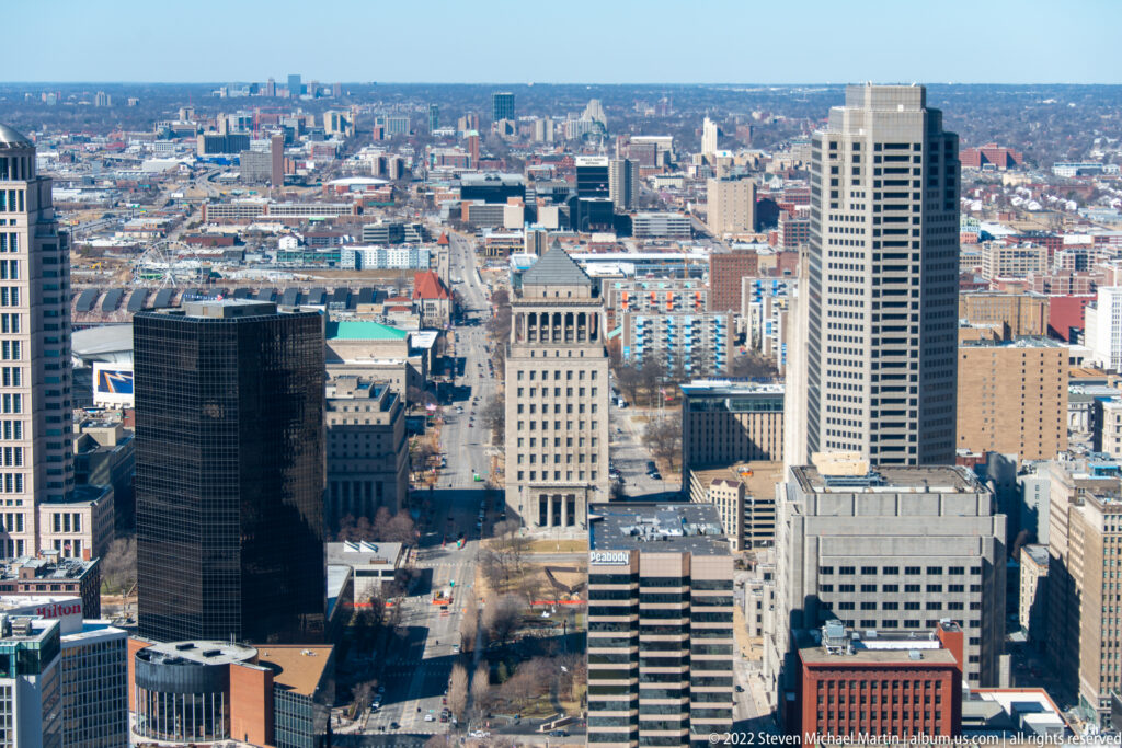 A high view of downtown St. Louis shows a dense skyline with dark and pale towers beneath a clear sky.