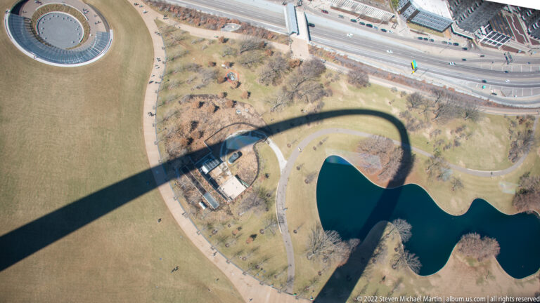 The Gateway Arch casts a long curved shadow across park paths and a reflecting pool in an aerial view.