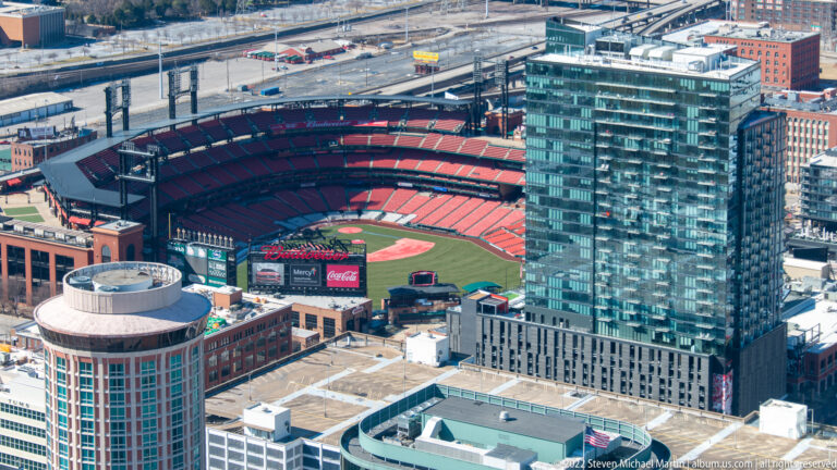 Busch Stadium’s red seating bowl and baseball field sit among downtown St. Louis buildings in a high view.