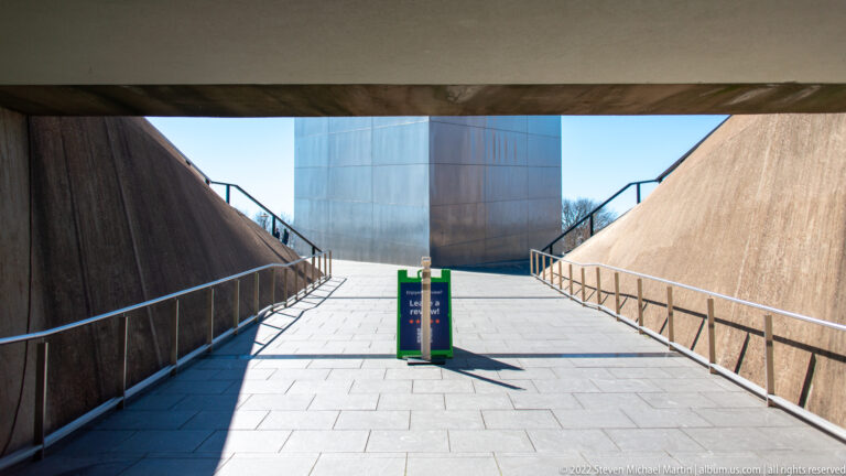 A paved walkway between sloped concrete walls leads toward the Gateway Arch base, with railings and a small sign.