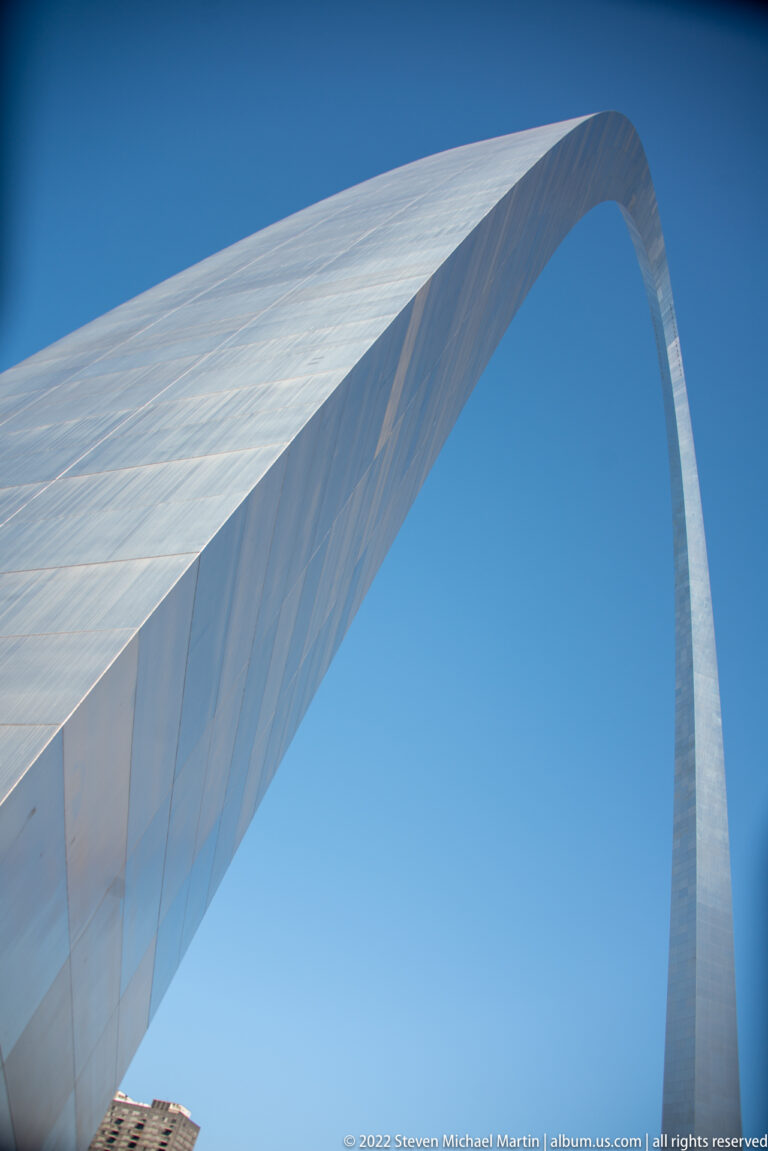 The stainless-steel Gateway Arch curves overhead against a clear blue sky, viewed from directly below.