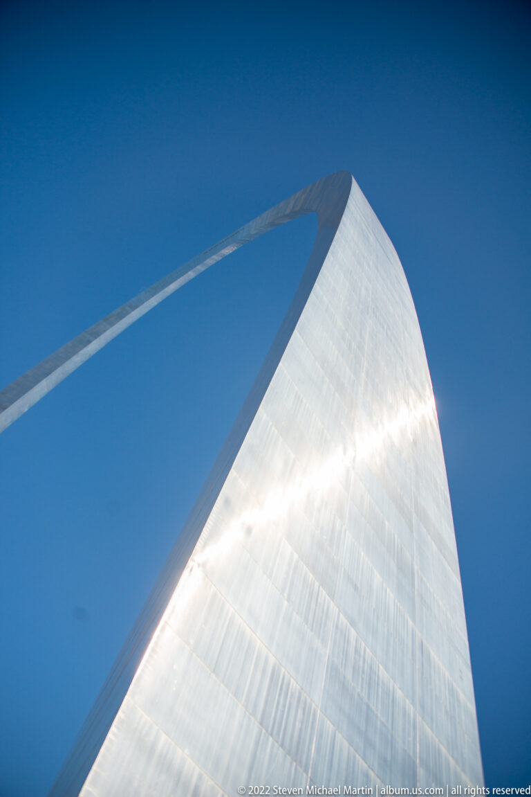 One reflective steel leg of the Gateway Arch rises overhead and joins the opposite leg against blue sky.