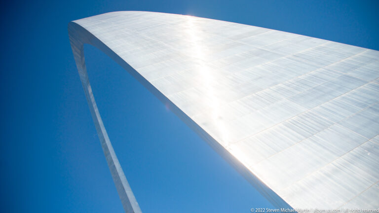 A close upward view shows the Gateway Arch’s reflective steel skin, triangular seam, and dark inner edge against blue sky.