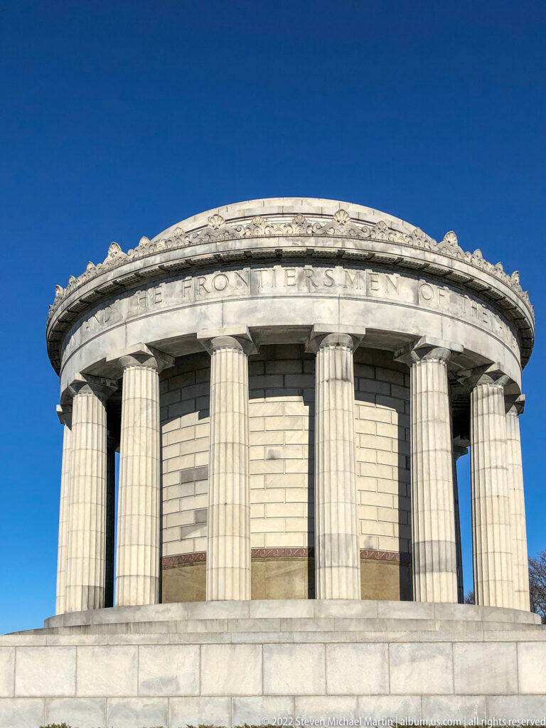 A curved limestone memorial façade with fluted columns and carved lettering rises against blue sky.