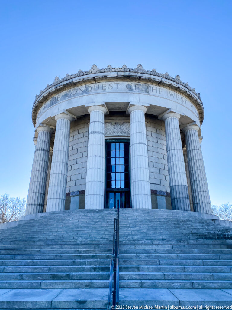 A centered stairway rises to the memorial’s dark doorway beneath a curved inscription.