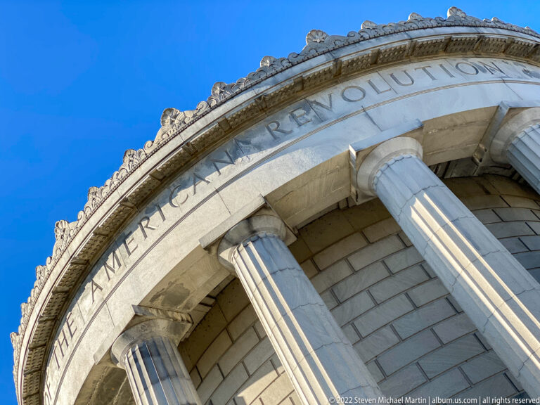 The words “The American Revolution” curve across the memorial’s stone frieze above columns.