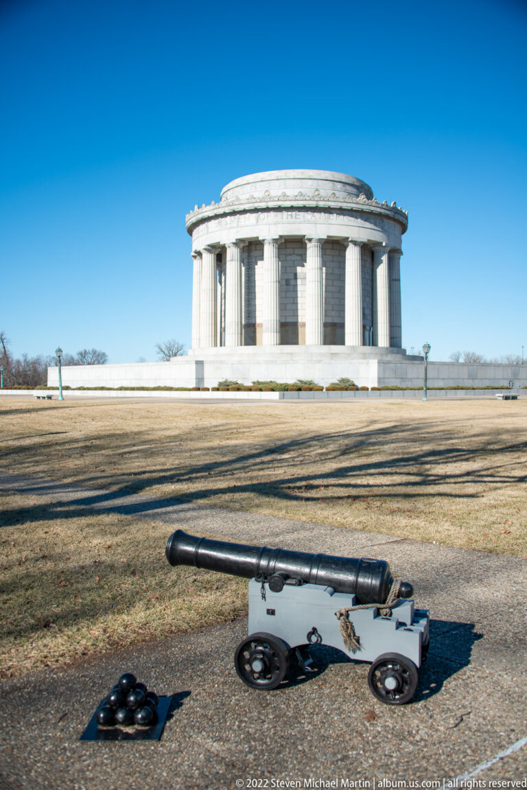 A cannon and cannonballs sit on the lawn with the memorial in the distance.