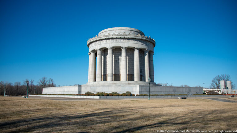 The round limestone memorial stands across an open lawn beneath a clear blue sky.