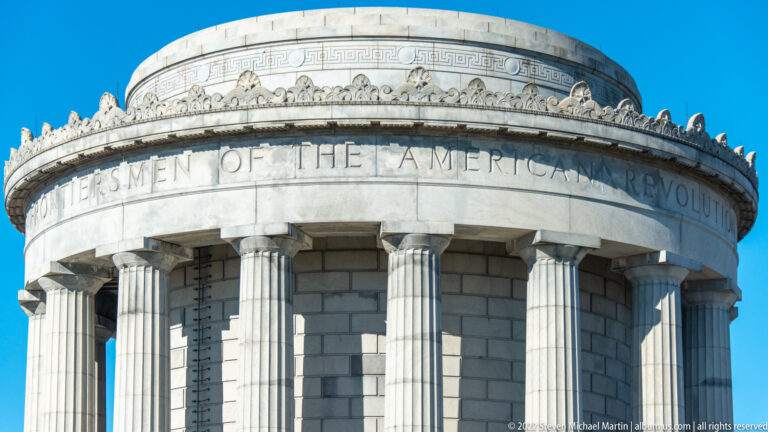 Carved lettering and ornamental stonework wrap around the memorial’s upper drum beneath a bright blue sky.