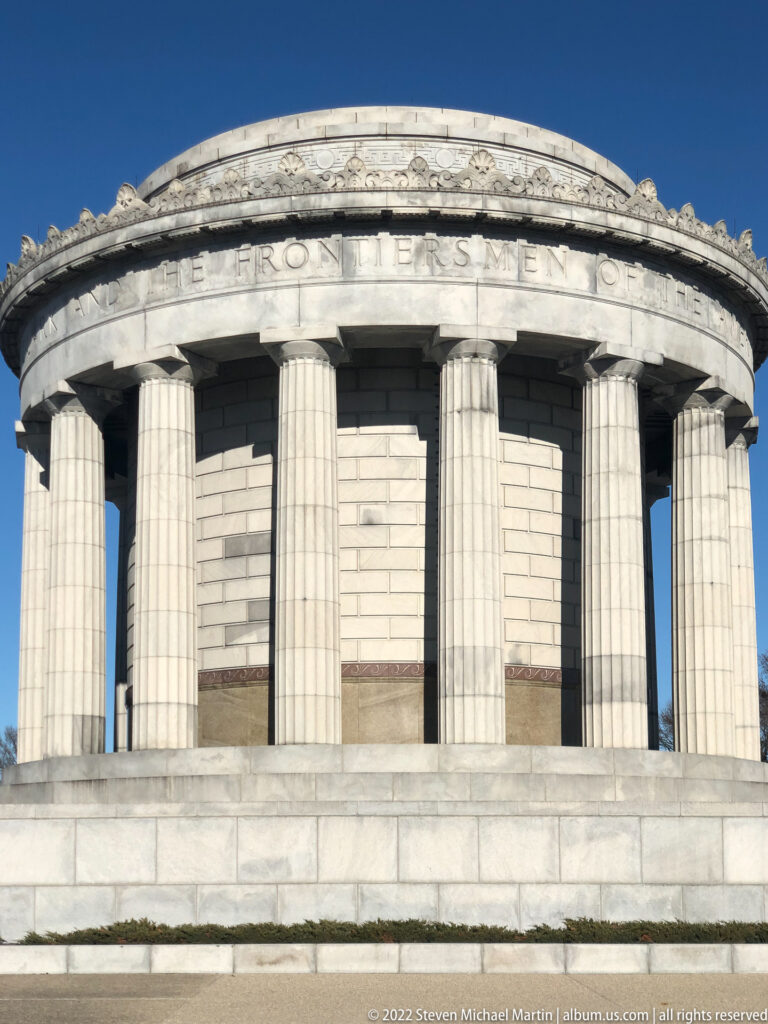 Pale stone columns stand beneath the memorial’s curved inscribed frieze, seen close against clear blue sky.