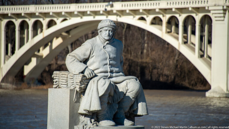 A seated stone figure in a cap and coat appears before an arched bridge over water.