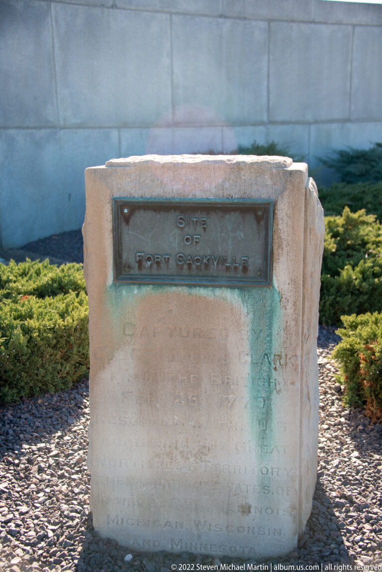 A small stone marker with an unreadable plaque stands among gravel and low shrubs.