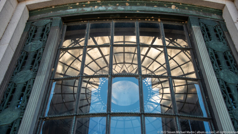 A gridded window reflects a circular pattern and sky beside ornamental metal panels.