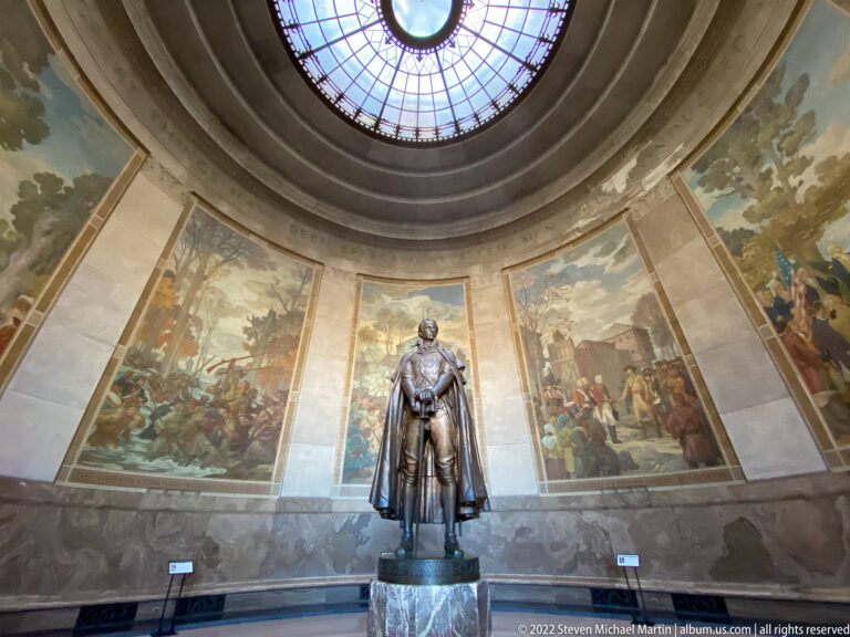 A bronze standing figure appears beneath a glass dome, surrounded by large historical murals.