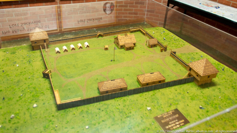 Elevated view of a miniature timber fort with blockhouses and small buildings arranged around an open yard.
