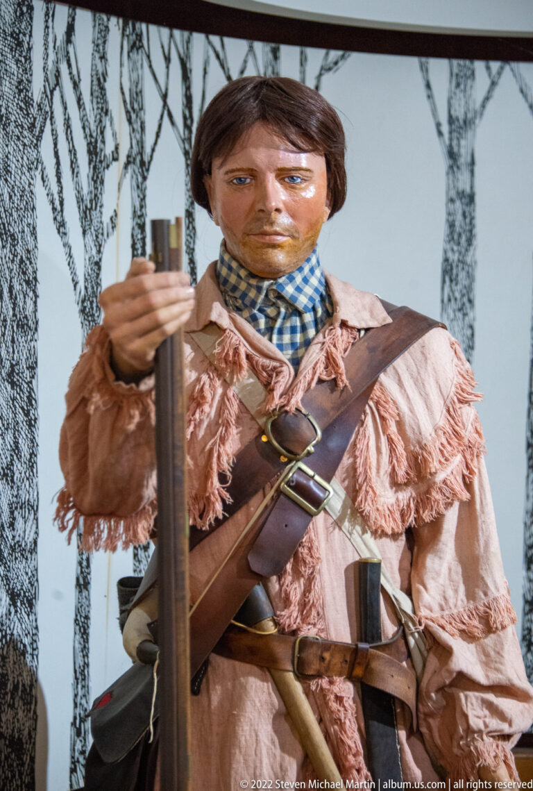 Mannequin in a fringed shirt and leather straps holds a long firearm against a woodland backdrop.