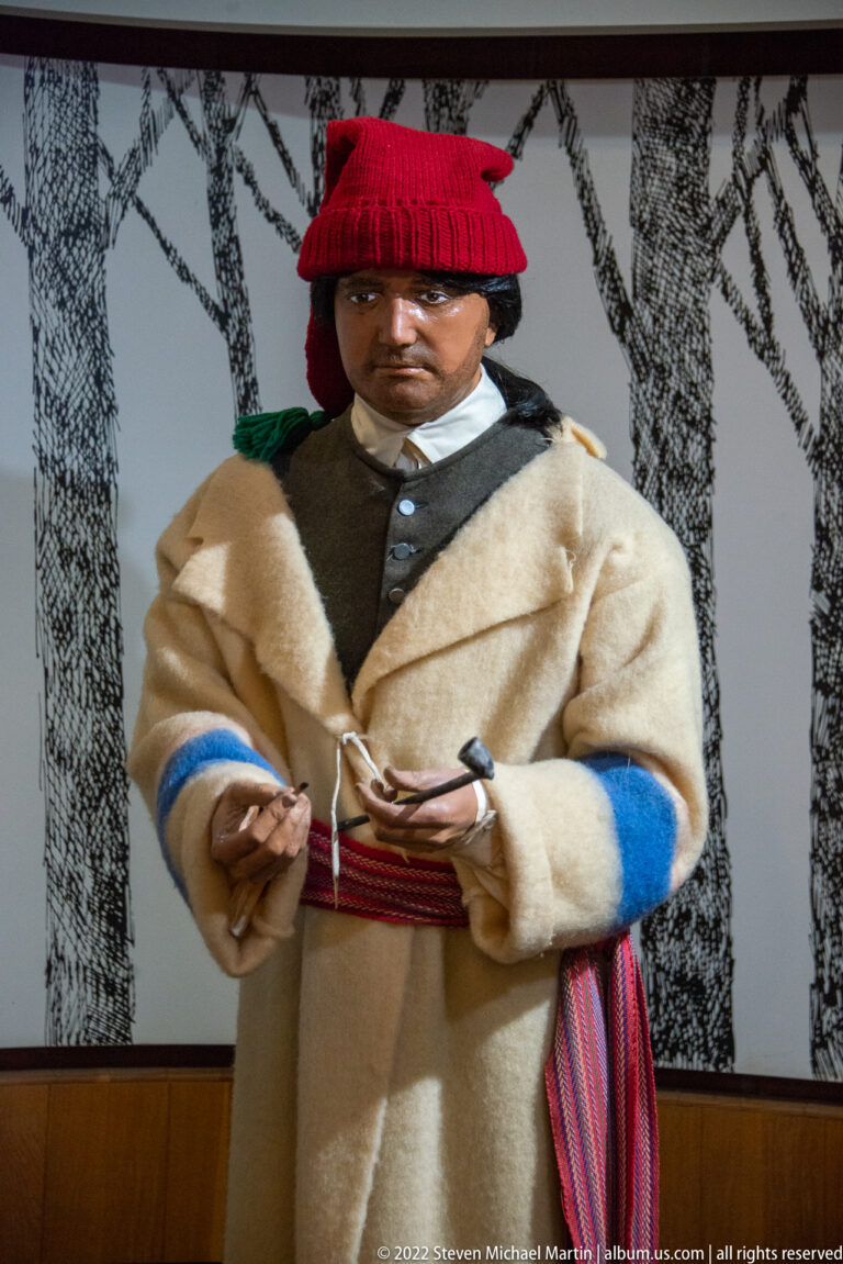 Mannequin in a red knit cap and pale wool coat with blue trim and a striped sash.