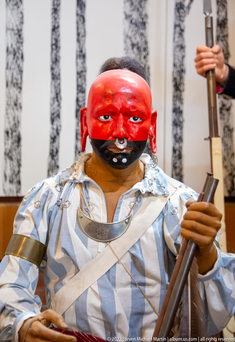 Seated mannequin with a red-painted face holds a long firearm in a patterned shirt and chest ornament.