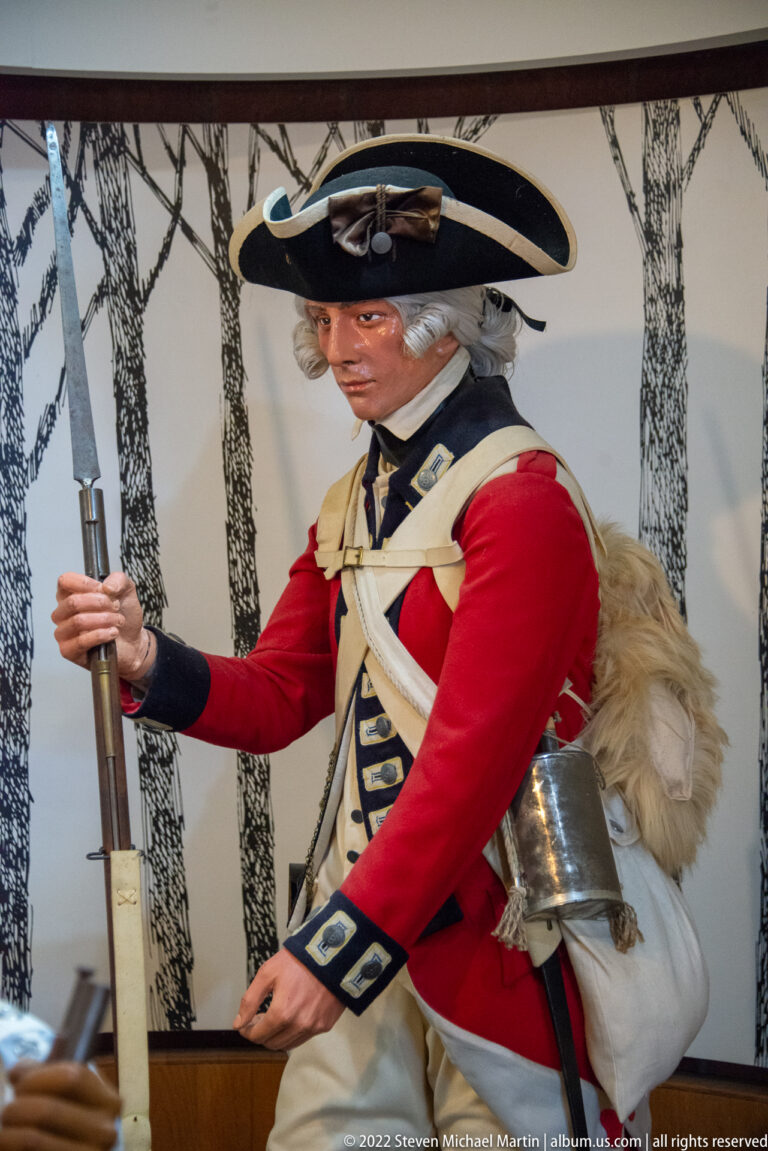 Red-coated mannequin in a tricorne hat leans forward while holding a musket.
