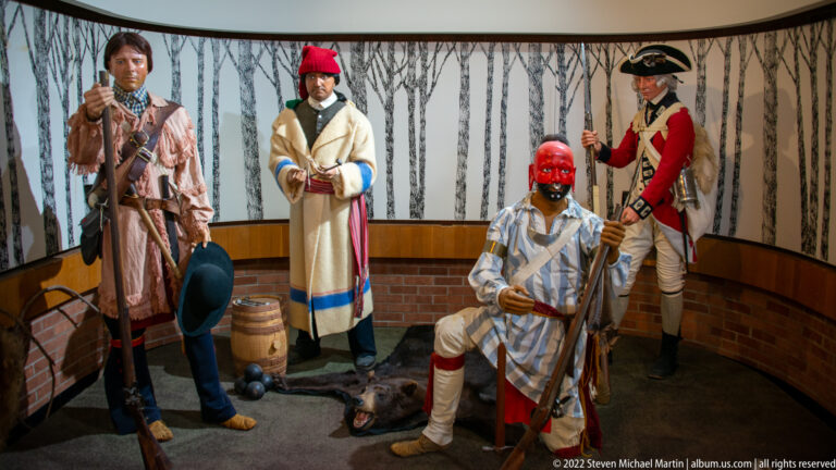 Several costumed mannequins stand among barrels and furnishings in a curved fort-themed exhibit room.
