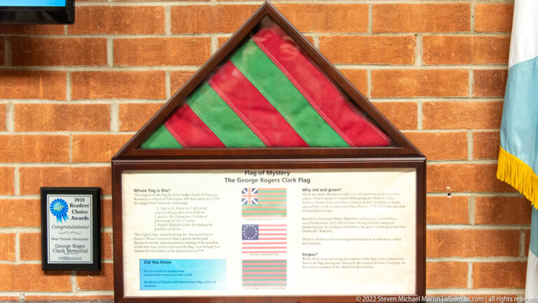 Red-and-green striped triangular flag above an interpretive panel on a brick wall.