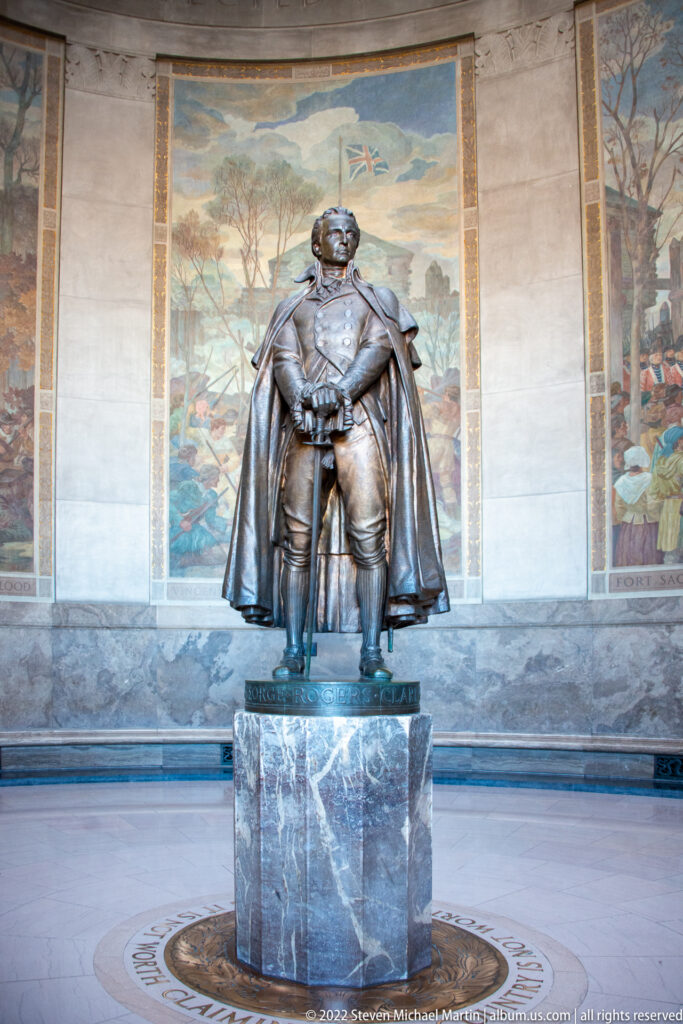 Bronze statue of a cloaked man on a marble pedestal before a large historical mural.