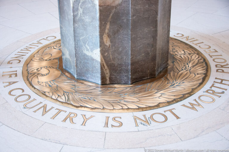 Circular floor medallion with a laurel wreath and lettering around a faceted marble pedestal.