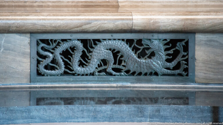 Close view of two intertwined dragon forms in a dark bronze grille set into marble.