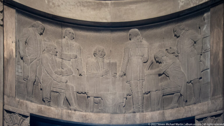 Pale carved relief of standing figures gathered around a seated person on a curved wall.