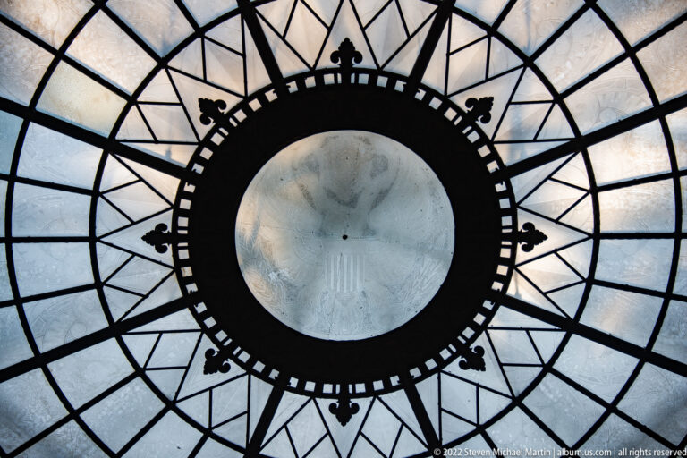 Upward view of a circular ceiling panel surrounded by radial dark metal ribs and glass.