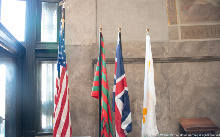 Four flags stand in a stone memorial chamber, with the United States flag at left and tall windows behind.