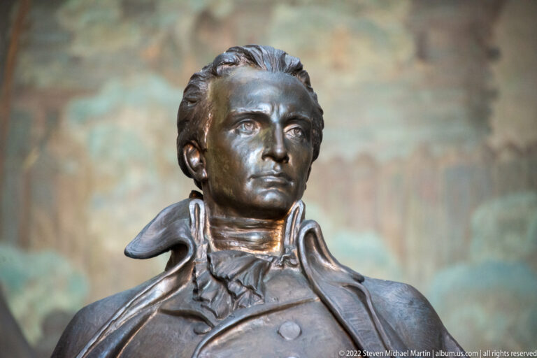 A bronze bust of George Rogers Clark shows him in a high-collared coat against a softly blurred stone background.
