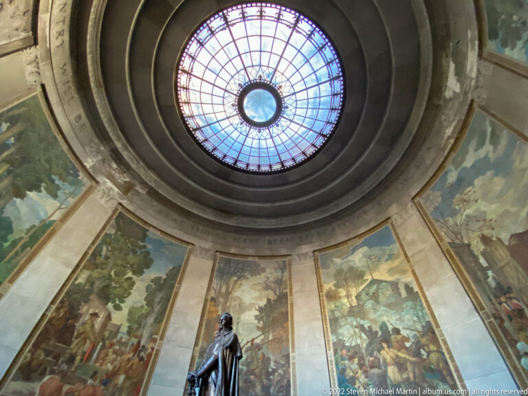 A small bronze figure stands below the rotunda’s glass dome and encircling murals.