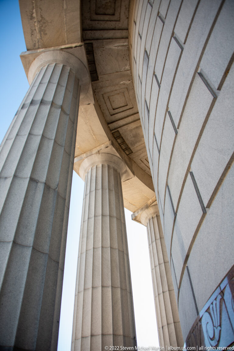Stone columns and a curved wall meet beneath a coffered memorial ceiling, photographed from a steep upward angle.